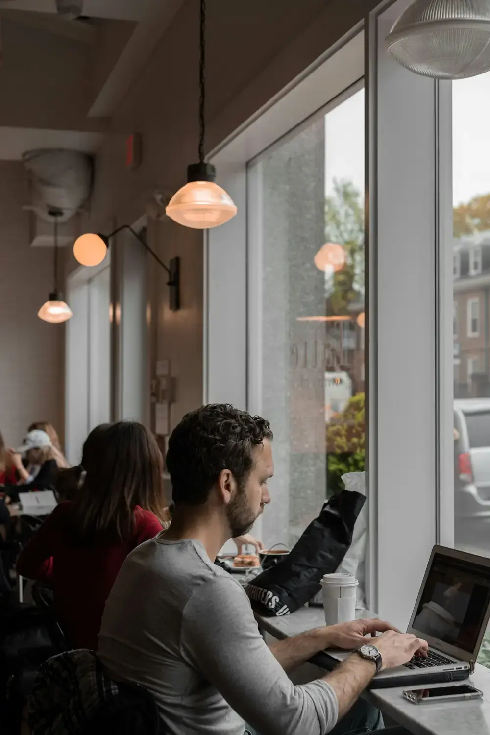 A man working on a laptop at a long window table in a cafe, with pendant lights behind him